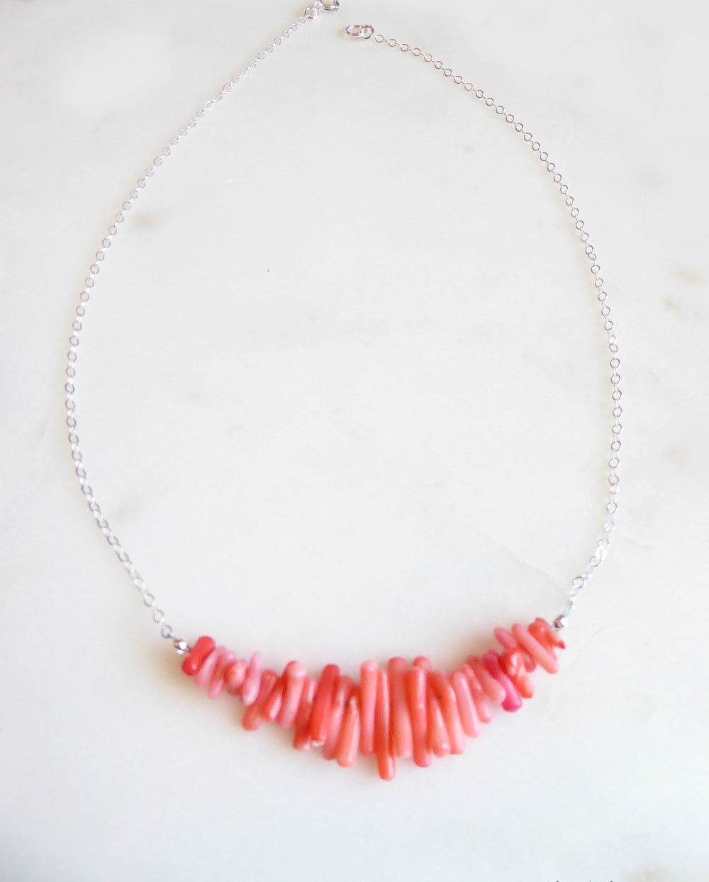 A necklace with a curved bar of irregular coral-pink beads on a delicate silver chain, displayed on a light marble surface.