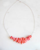 A necklace with a curved bar of irregular coral-pink beads on a delicate silver chain, displayed on a light marble surface.