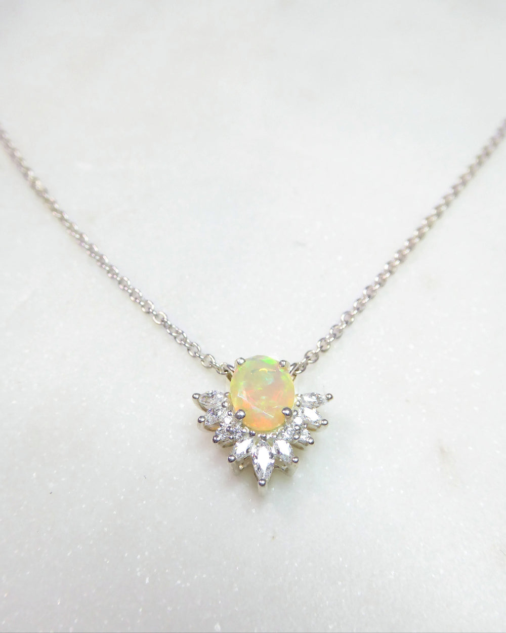 A delicate silver chain necklace featuring a central oval opal pendant surrounded by small clear stones, displayed on a light marble surface. The brand name 'Glass Palace Arts' is printed in the upper background.