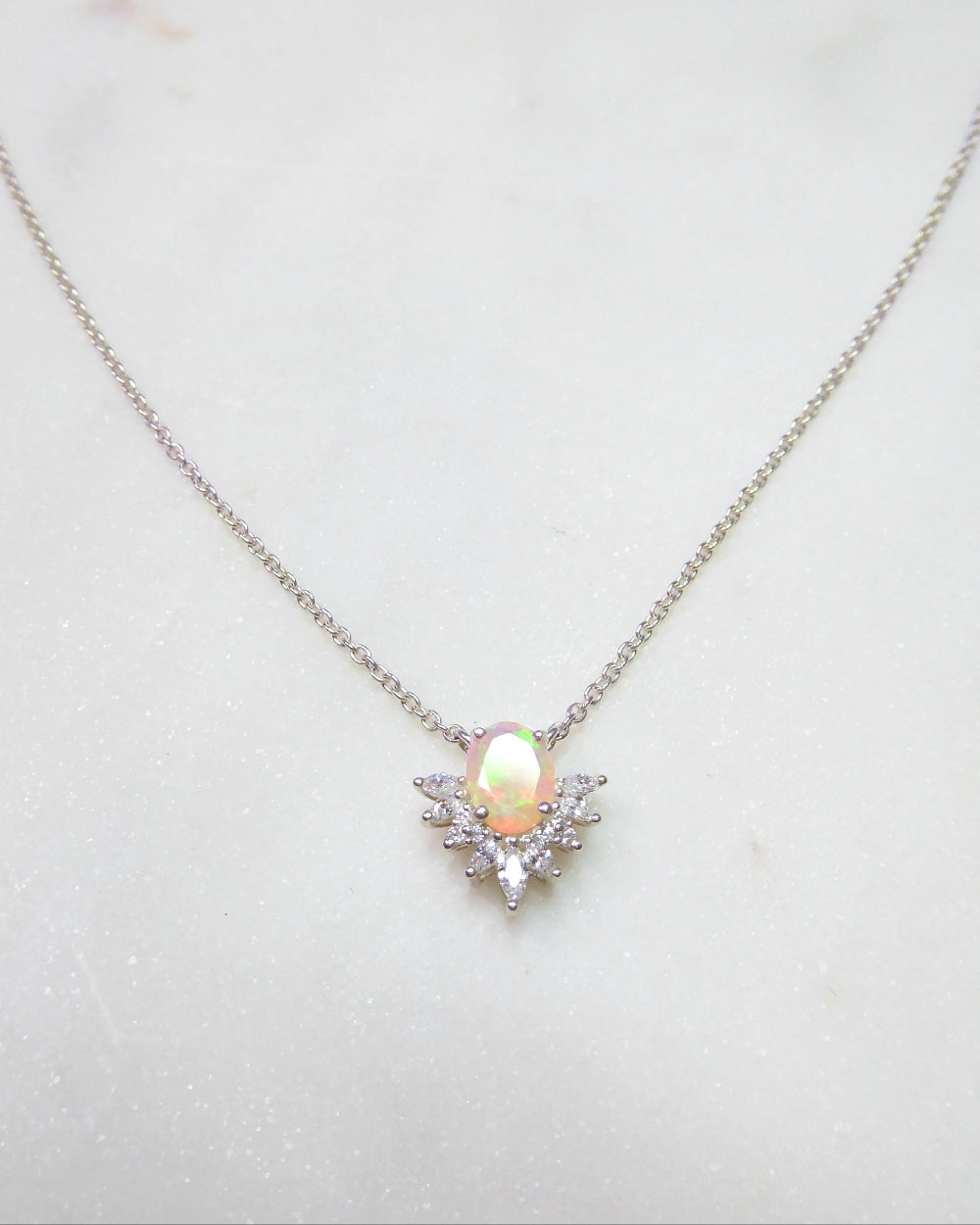 A delicate silver chain necklace featuring a central pendant with a pear-shaped iridescent opal gemstone surrounded by small clear crystals, displayed on a light marble surface.